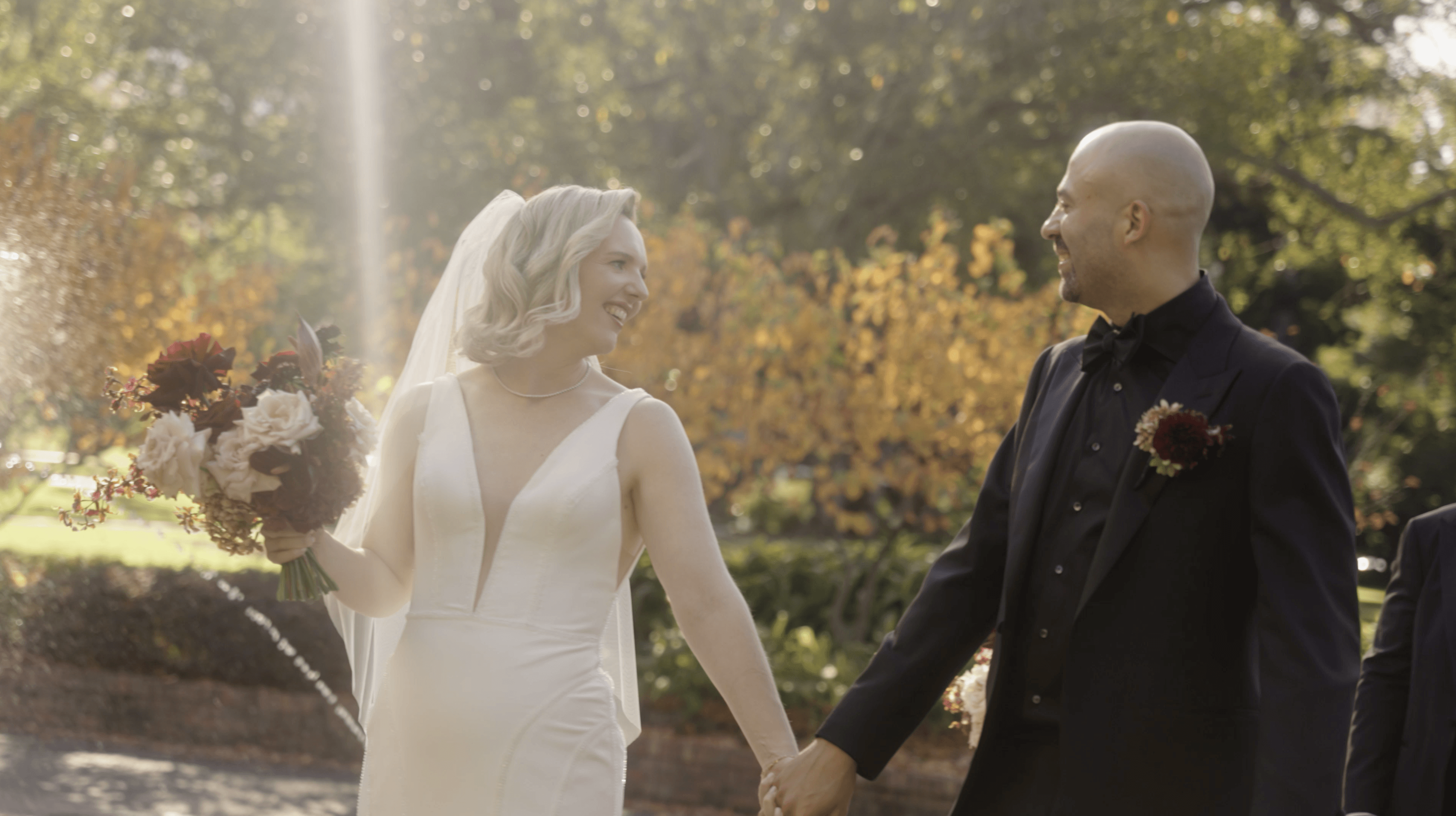 A bride and groom pour champagne into a tiered cascade of glasses, adorned with purple grapes, during a festive outdoor celebration.