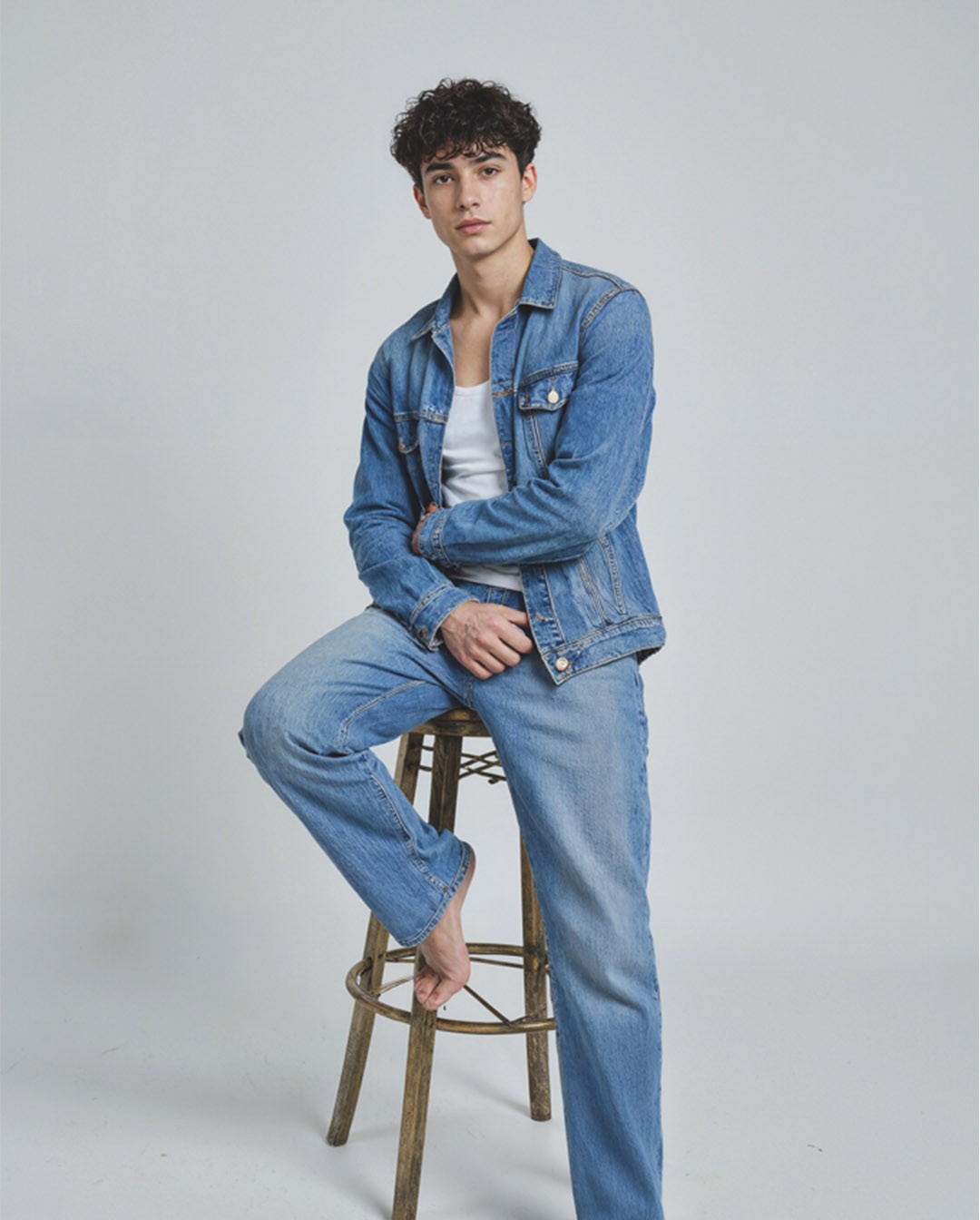 Young man sitting on a wooden stool wearing a blue denim jacket and jeans with a white t-shirt, casual studio fashion portrait