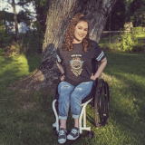 A young woman with curly hair smiles while sitting in a wheelchair outdoors, next to a large tree. She is wearing a dark t-shirt, jeans, and sneakers.