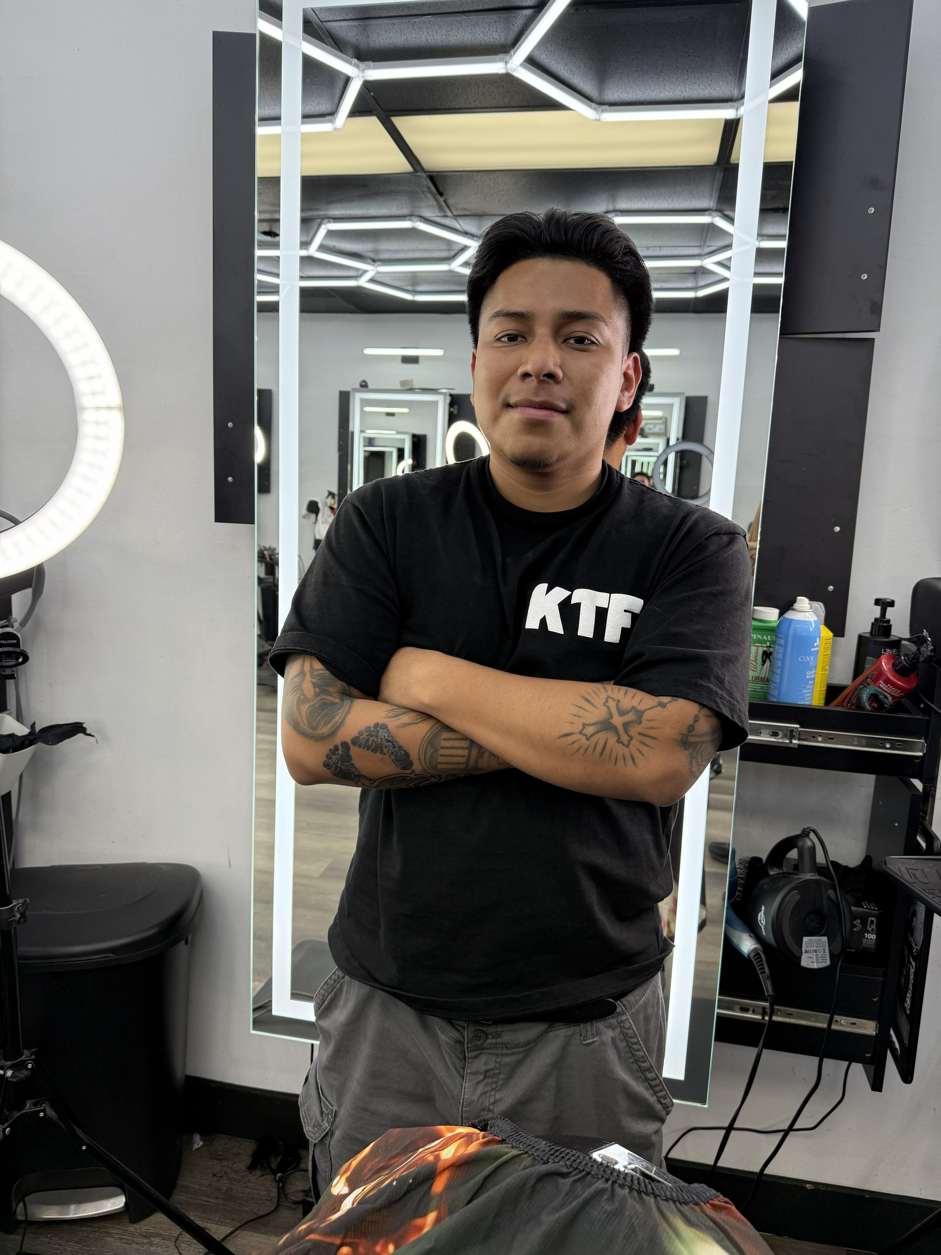 A barber shapes a client’s haircut using clippers near a bright ring light in barbershop. 
