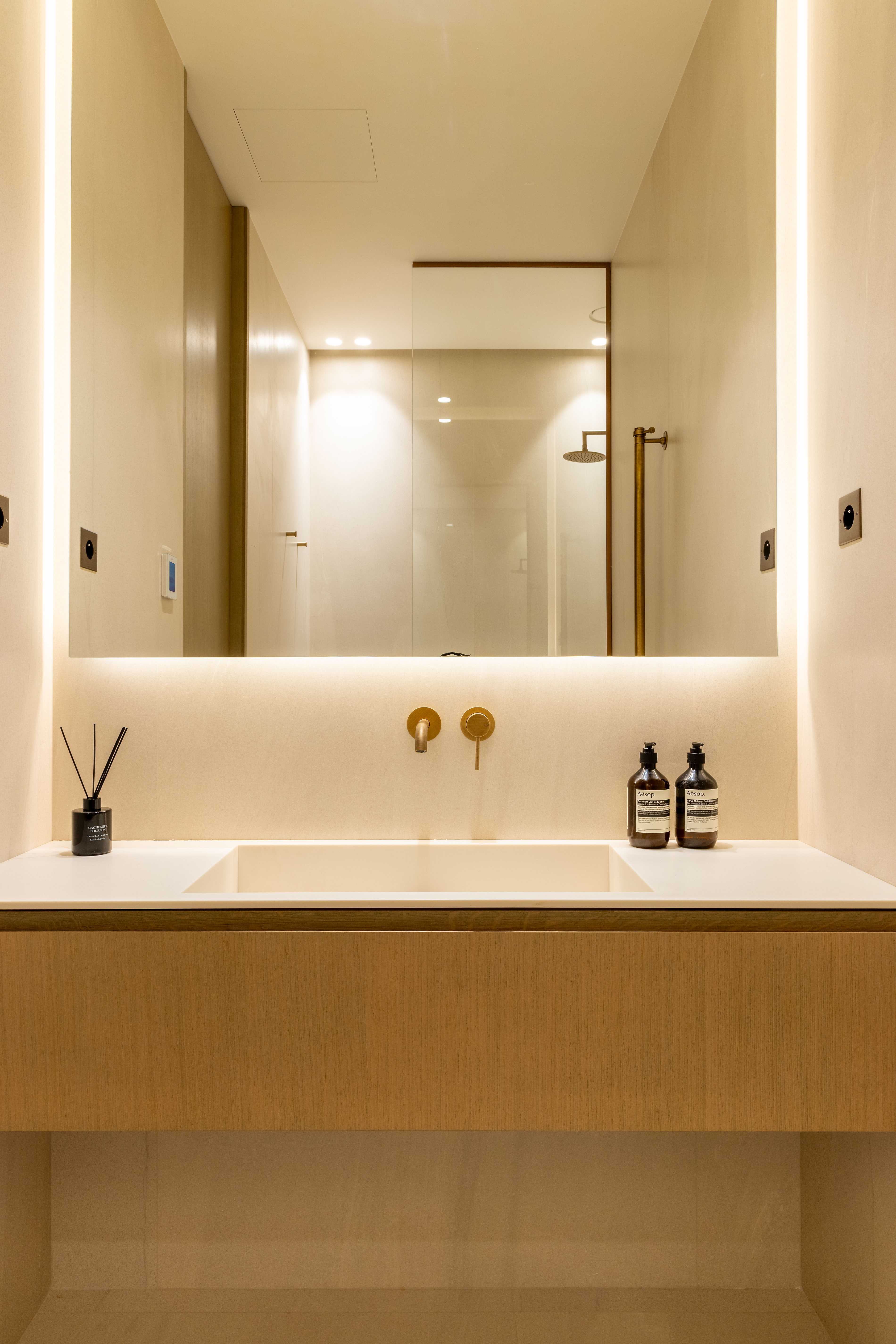Vasque avec grand miroir rétroéclairé, robinetterie dorée, produits Aesop et reflet de la douche