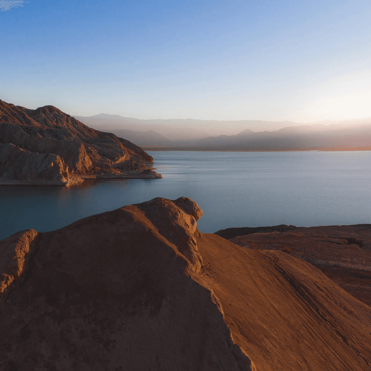 Sunset over a serene lake with rugged, sunlit brown cliffs in the foreground and distant mountains under a clear blue sky. Calm and peaceful atmosphere.