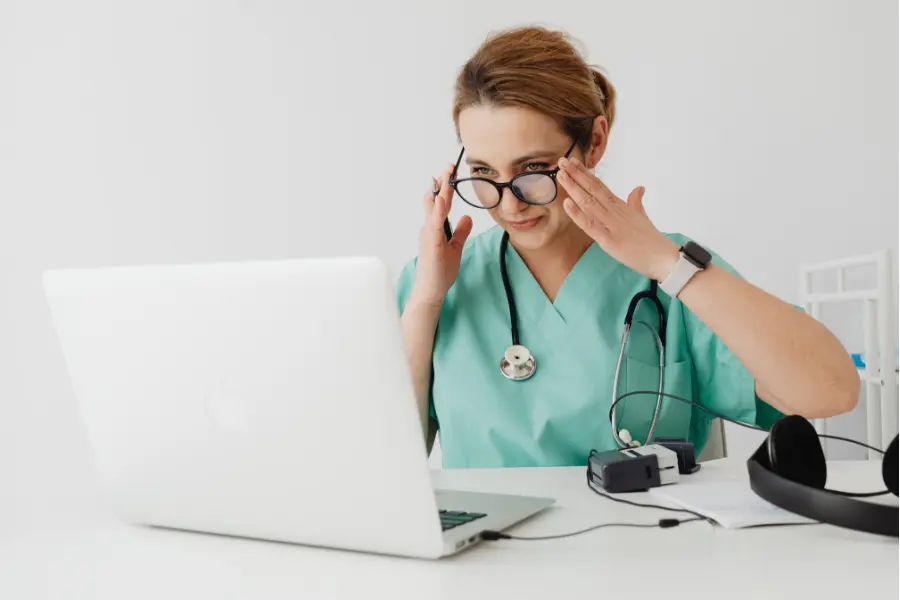 AI admin assistant for healthcare shown as a nurse in green scrubs adjusting her glasses while looking at a laptop.