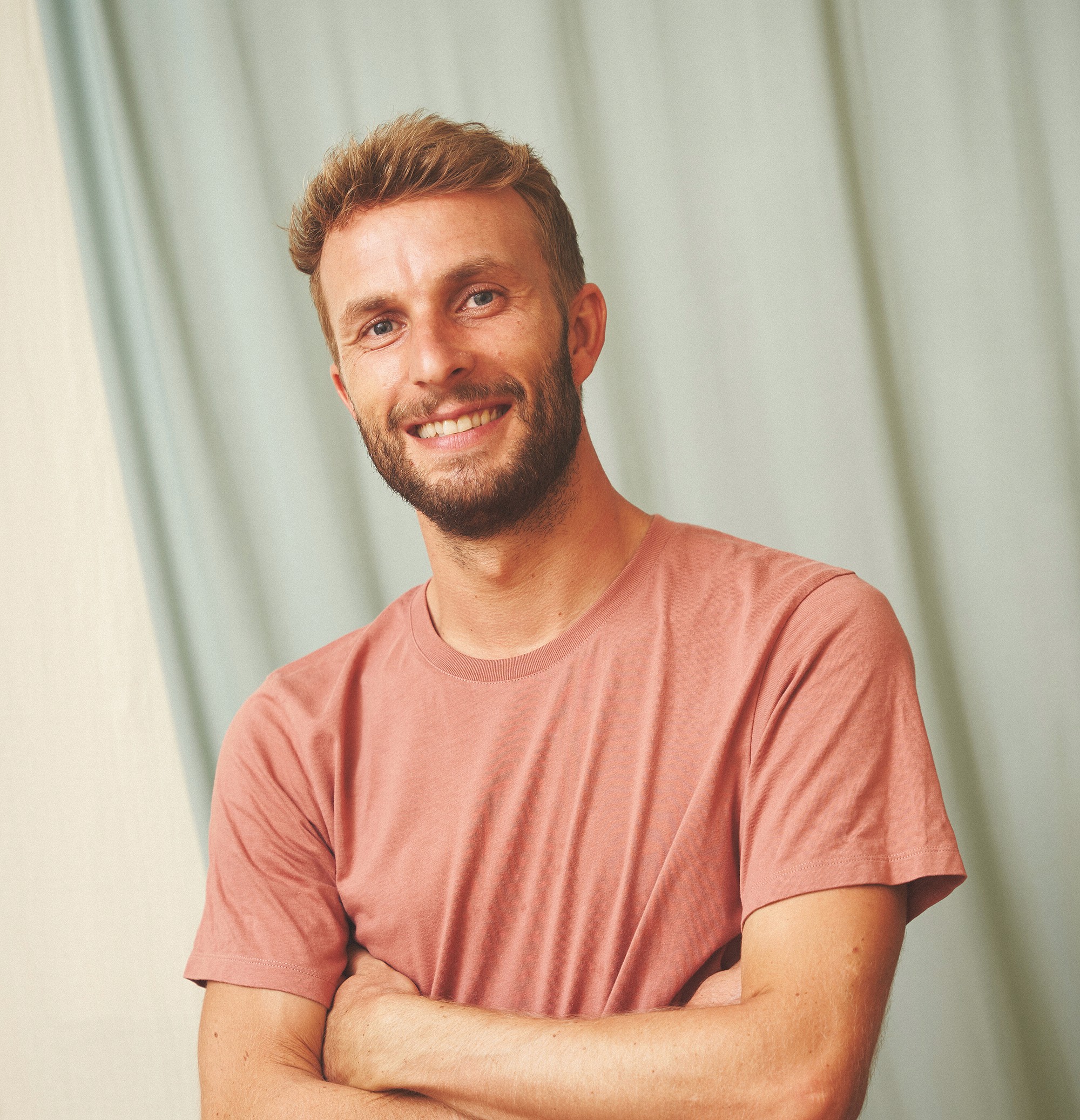 Person wearing a blue puffer jacket and red backpack standing in front of a light blue building on a city street.