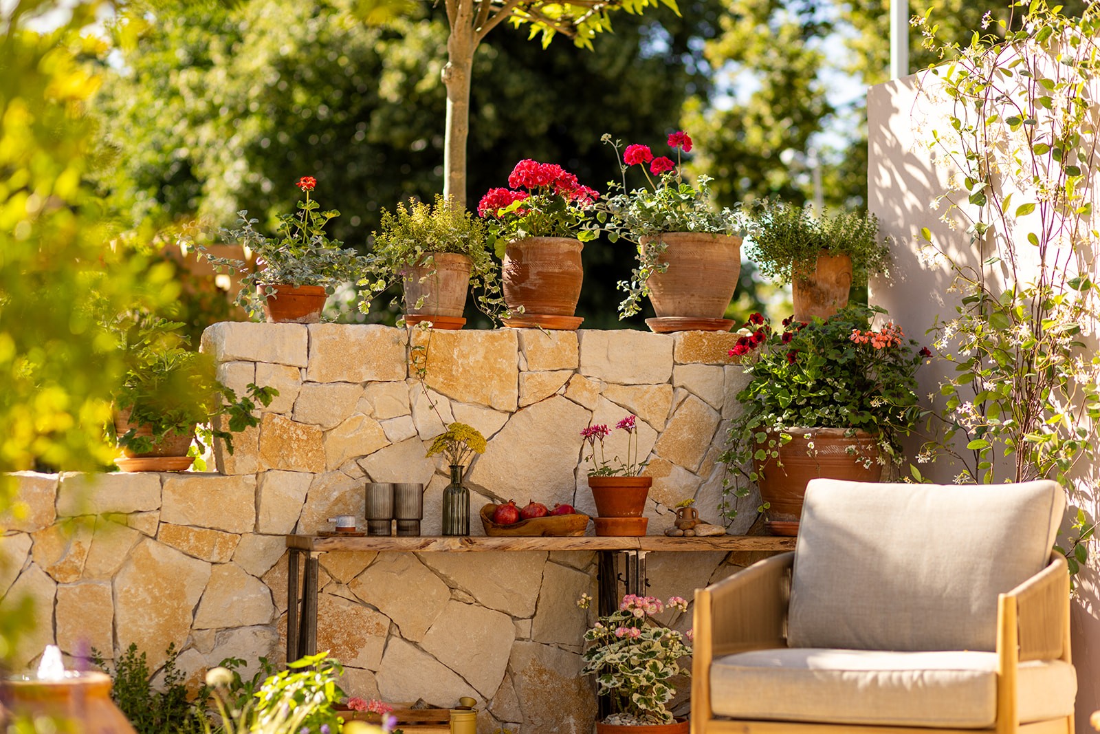 A cozy garden scene featuring potted plants, a stone wall, and a comfortable chair under dappled sunlight.