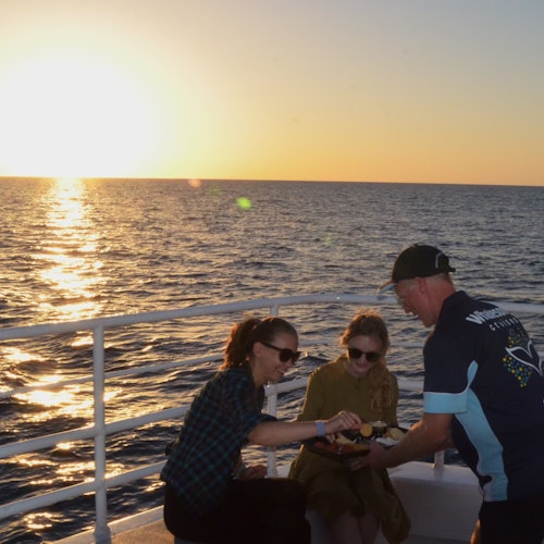 Two women sitting on a boat at sunset, with another person handing them a plate of food. The ocean and sun are visible in the background.