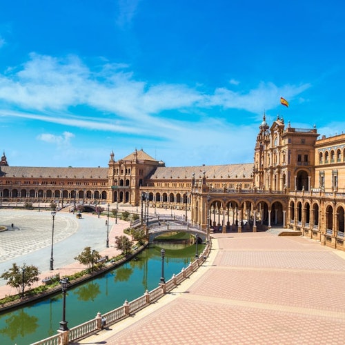 Amplia plaza con edificios arqueados, una vía fluvial central y una bandera izada, todo bajo un cielo azul brillante.