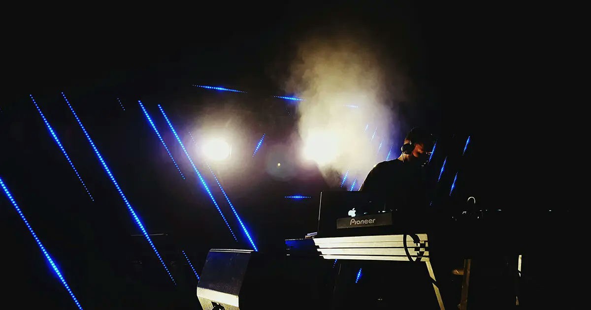 DJ performing on stage with moody lighting, blue LED visuals, and atmospheric fog.