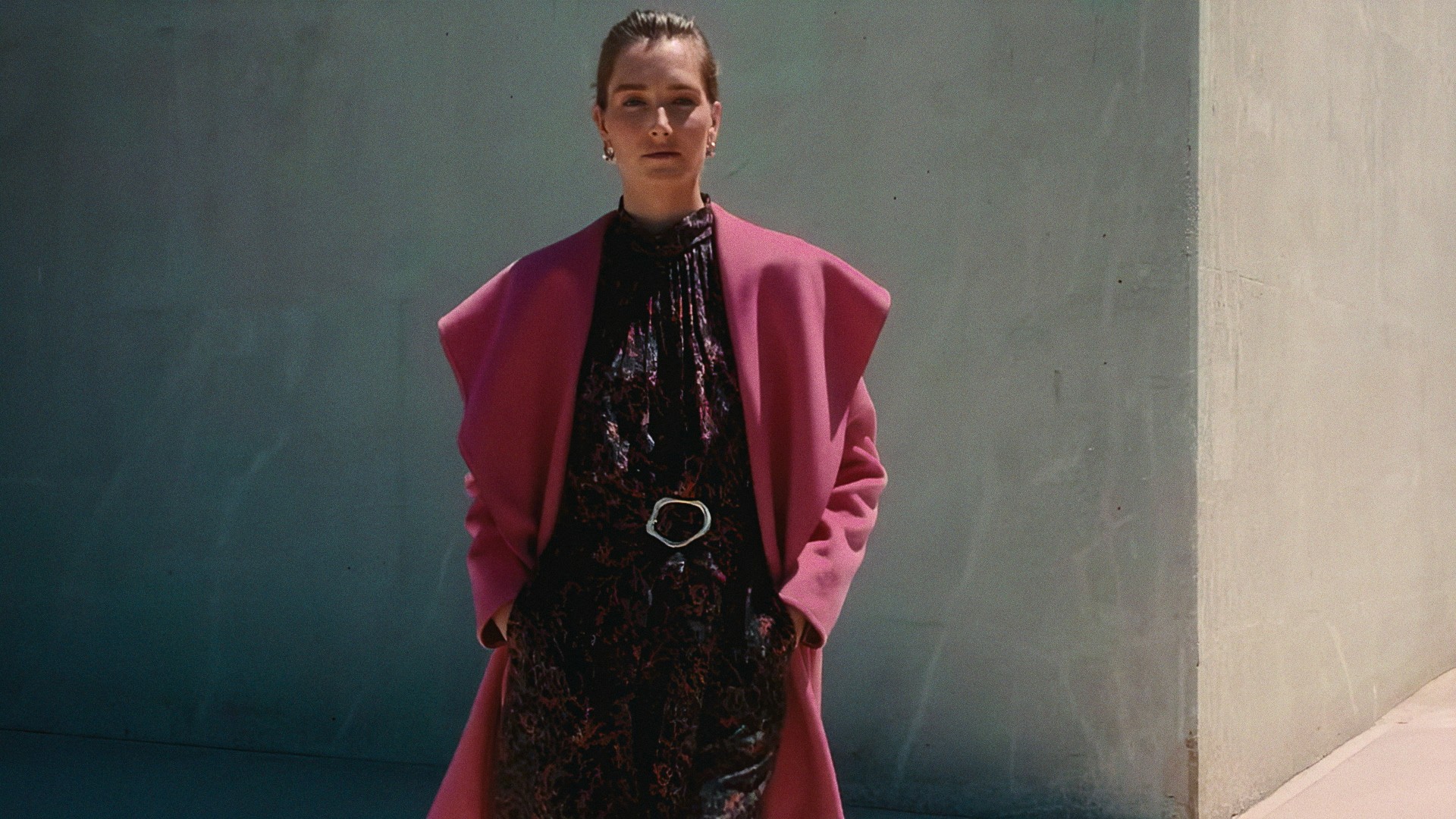 A woman in a pink coat over a black printed dress against a gray concrete wall, Lafayette 148 New York