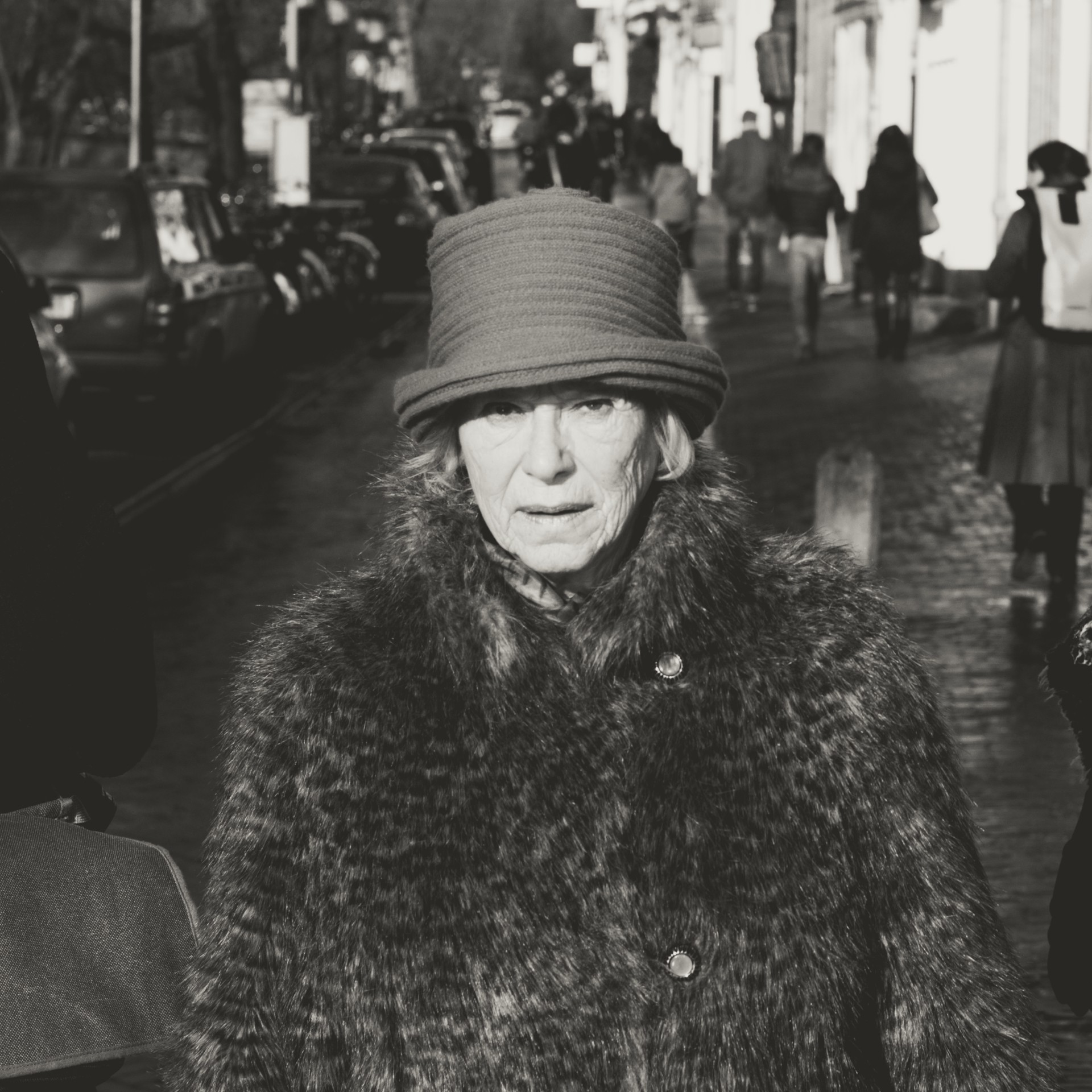 Elderly woman in fur coat and hat standing on city street with cars and pedestrians.
