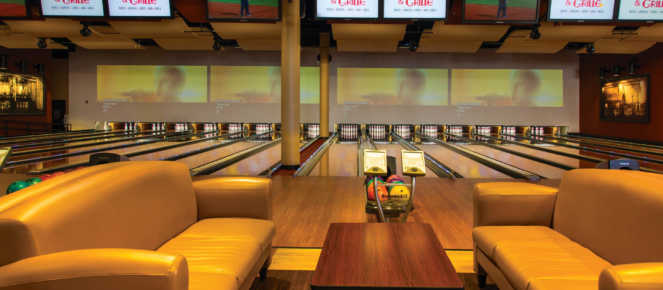 A young person with curly hair smiles at the camera while sitting in front of a laptop against a dark background.