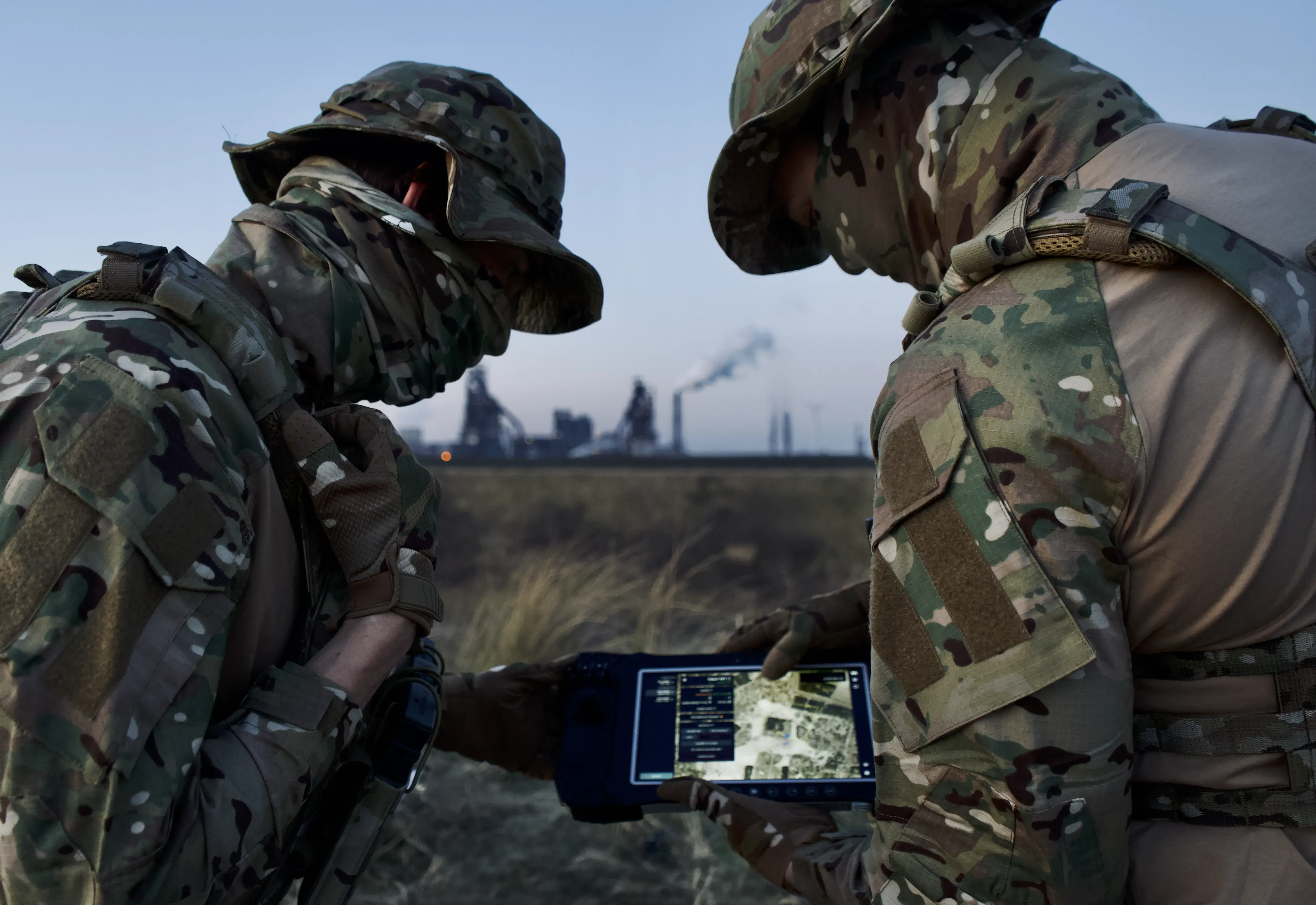 Two soldiers in camouflage uniforms are analyzing a digital map on a tablet in an open field, with smoke-emitting industrial structures visible in the distant background.