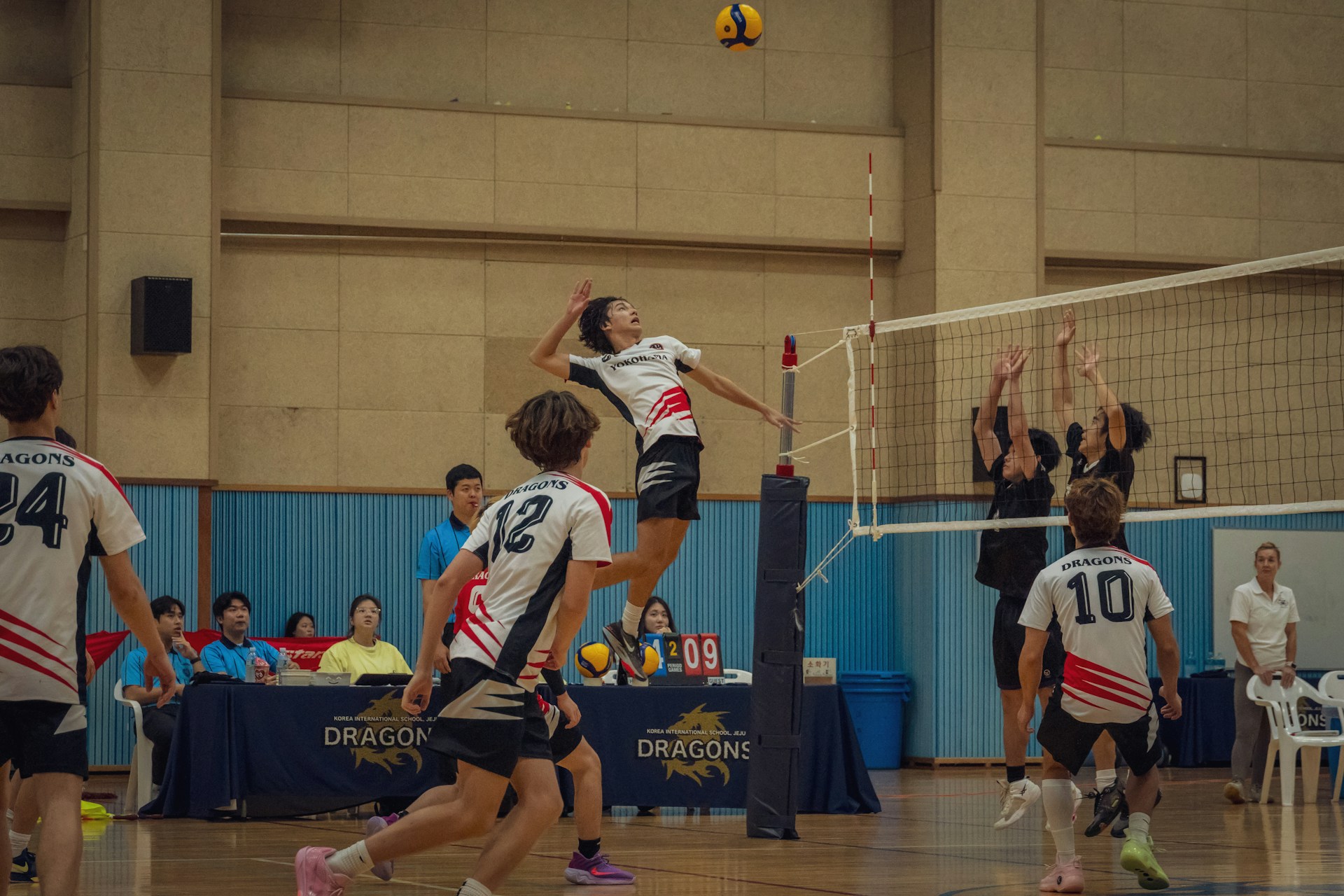 A volleyball player in a white and red jersey jumps high to spike the ball over the net against opposing blockers.