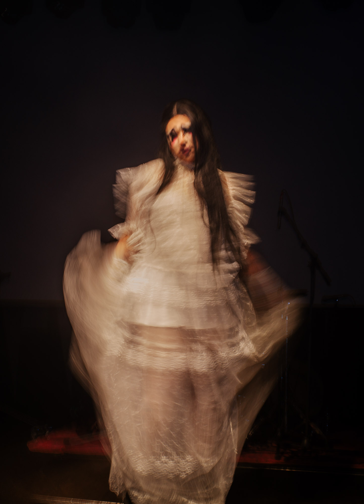 A person is captured in motion wearing a flowing, translucent white dress with ruffled details, blurred to convey movement against a dimly lit background, suggesting a dramatic or theatrical performance.