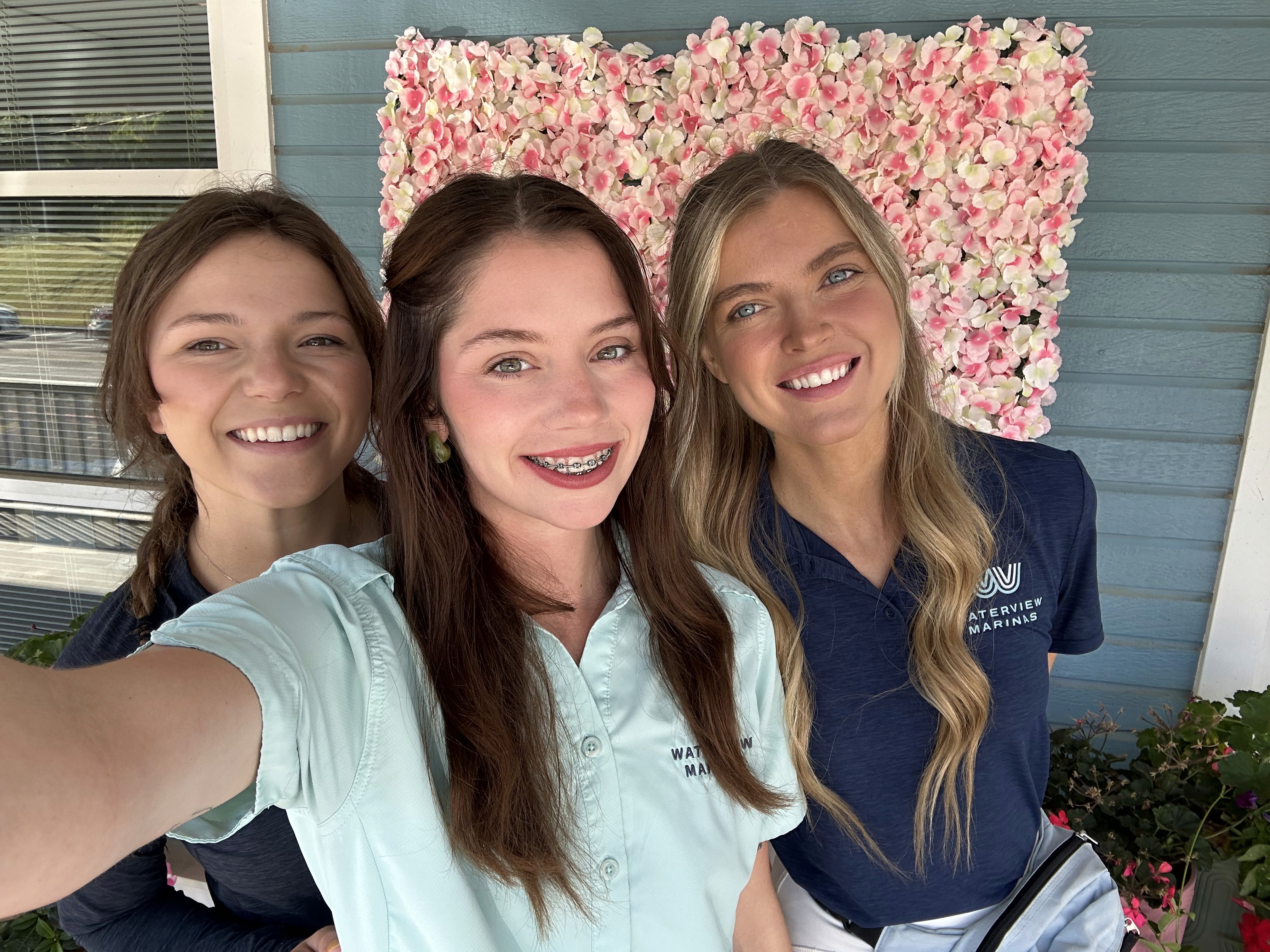 Three smiling individuals stand closely together in front of a decorative pink flower wall, with two wearing embroidered shirts featuring a wave design logo, set against a blue building exterior.