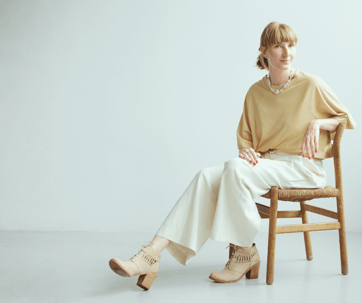 A woman with a short hairstyle sits on a wooden chair in a minimalist setting. She wears a light yellow top, cream wide-leg pants, and beige chunky heels, exuding a relaxed and stylish vibe.