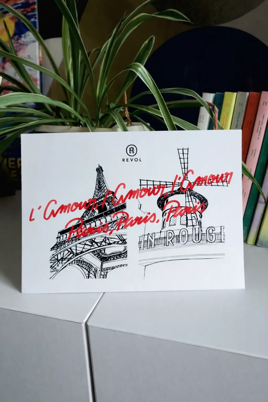 Carte blanche de la marque Revol posée sur un meuble. Elle présente des illustrations à l'encre noire de la Tour Eiffel et du Moulin Rouge, surmontées d'une inscription manuscrite rouge répétée : 'L'Amour, l'Amour, l'Amour, Paris, Paris, Paris'.