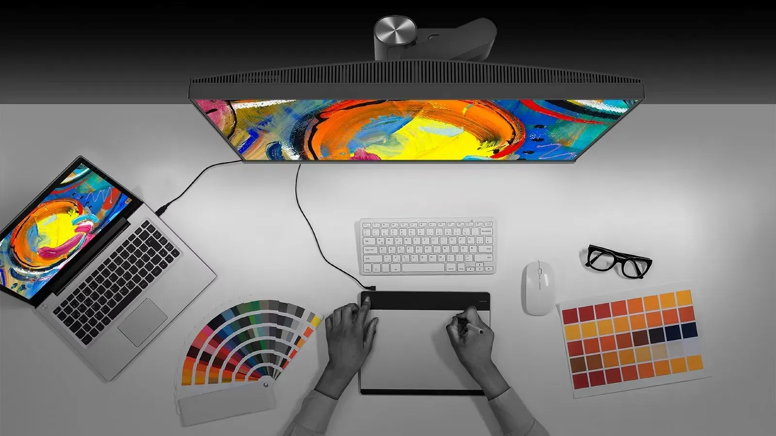 A person is using a digital drawing tablet connected to a computer, with colourful abstract art displayed on the monitor, surrounded by a laptop, colour swatches, and eyeglasses on a white desk.