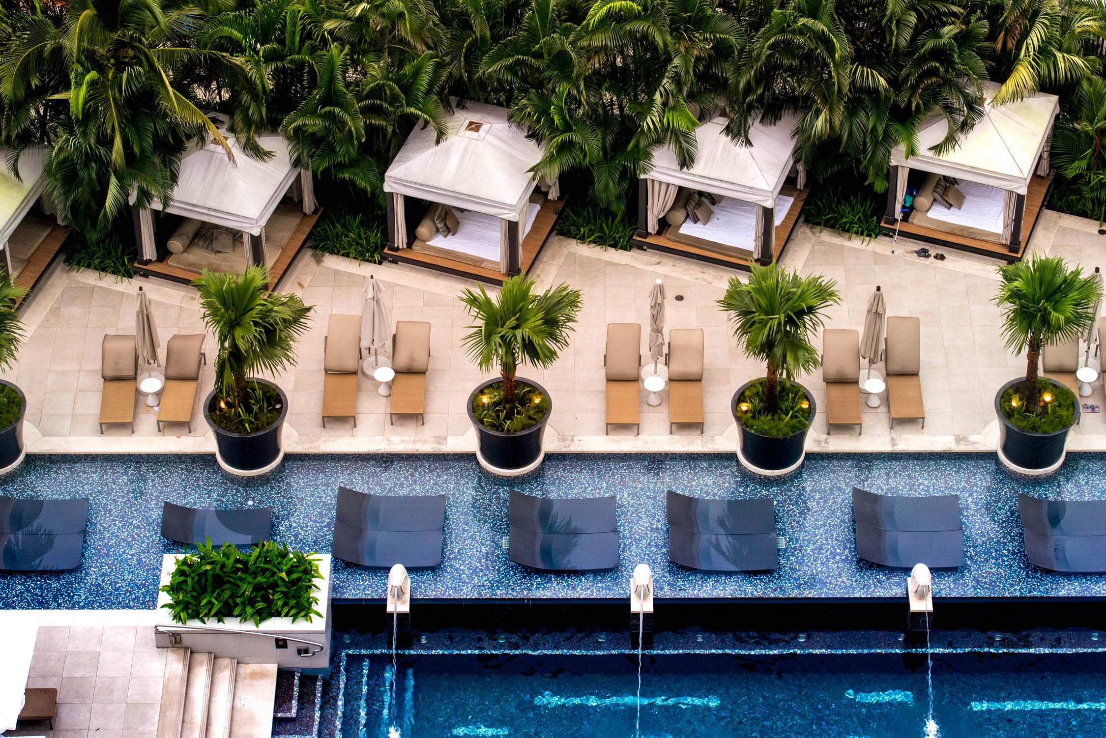 Lounge chairs and cabanas with white roofs line a pool deck shaded by lush green palm trees under bright daylight.