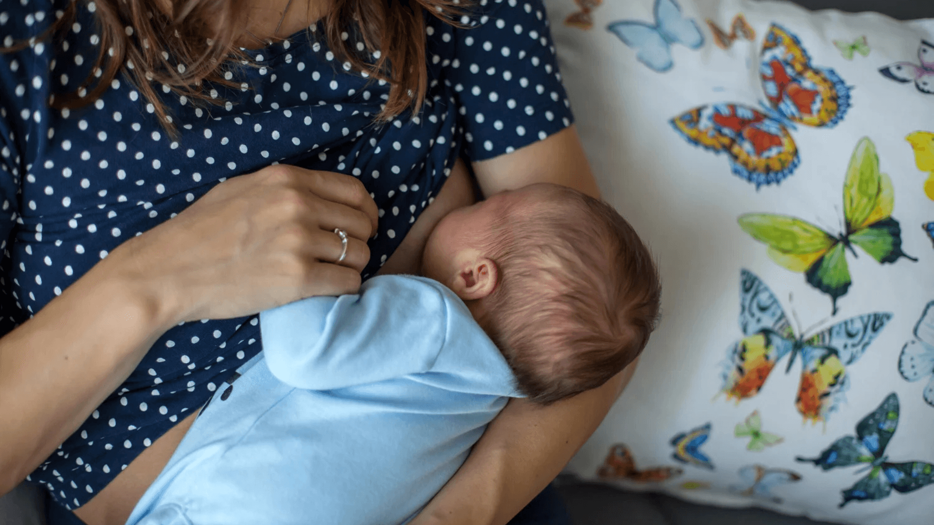 Mother breastfeeding a newborn baby while sitting beside a colorful pillow decorated with butterfly illustrations.