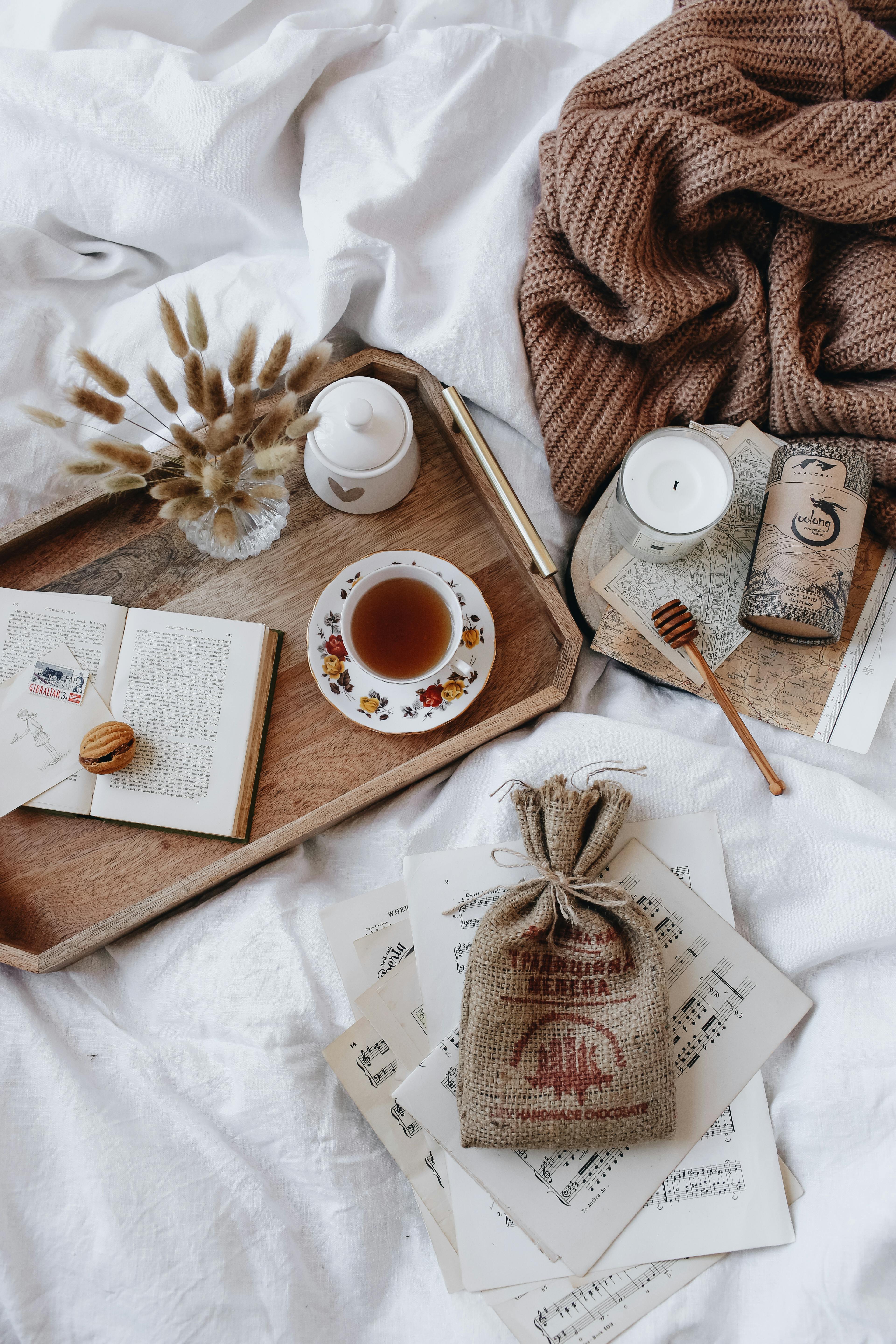 An aesthetic photo featuring a tray with a book, a cup of tea, and a candle, along with papers, a small pouch, and another candle placed beside the tray.