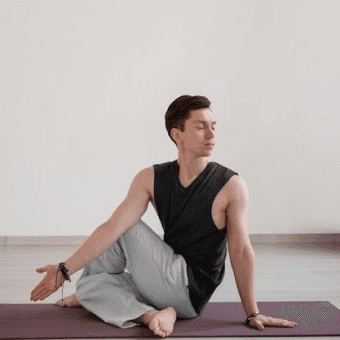 Man in a yoga twist pose on a purple mat, wearing a black tank top and light pants. Practicing yoga.