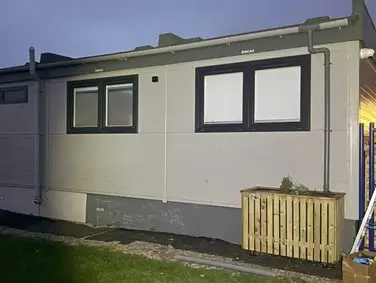 A side view of a simple modern house with two windows and a small outdoor unit in the foreground.