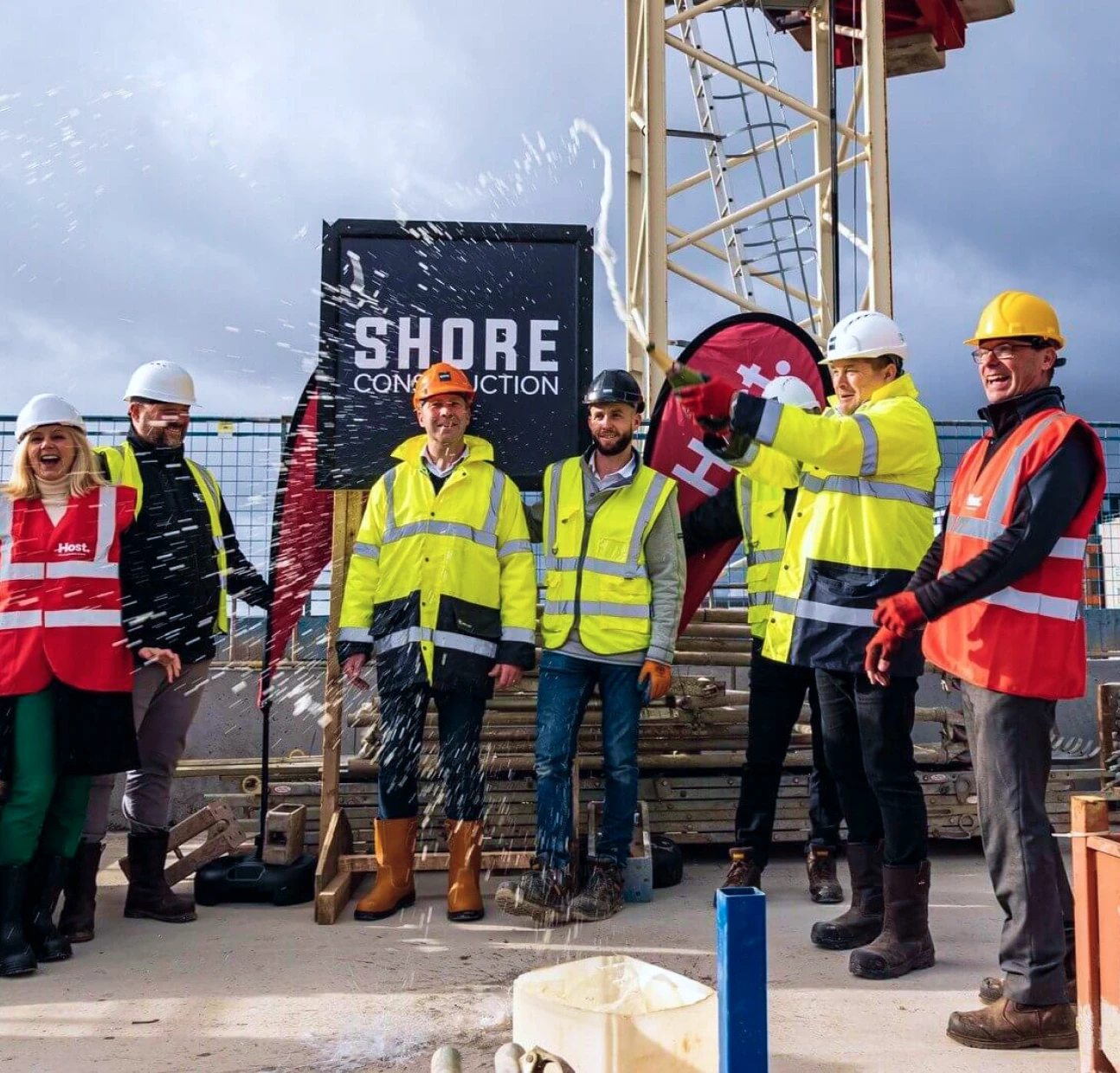 A group of construction workers in high-visibility jackets and helmets celebrate a groundbreaking ceremony at a construction site, with champagne being sprayed and a backdrop that includes a sign reading "Shore Construction" and construction equipment, under a cloudy sky.