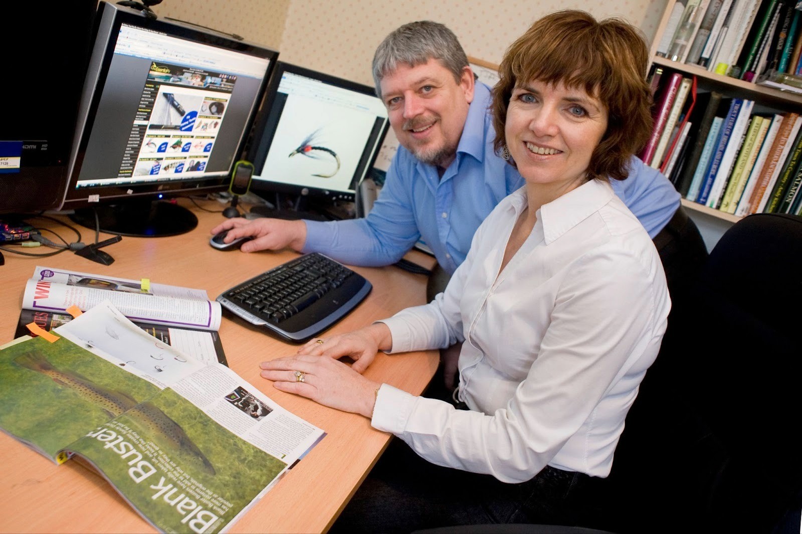 A man and woman sit at a desk, focused on computer screens, discussing fly fishing materials and techniques.