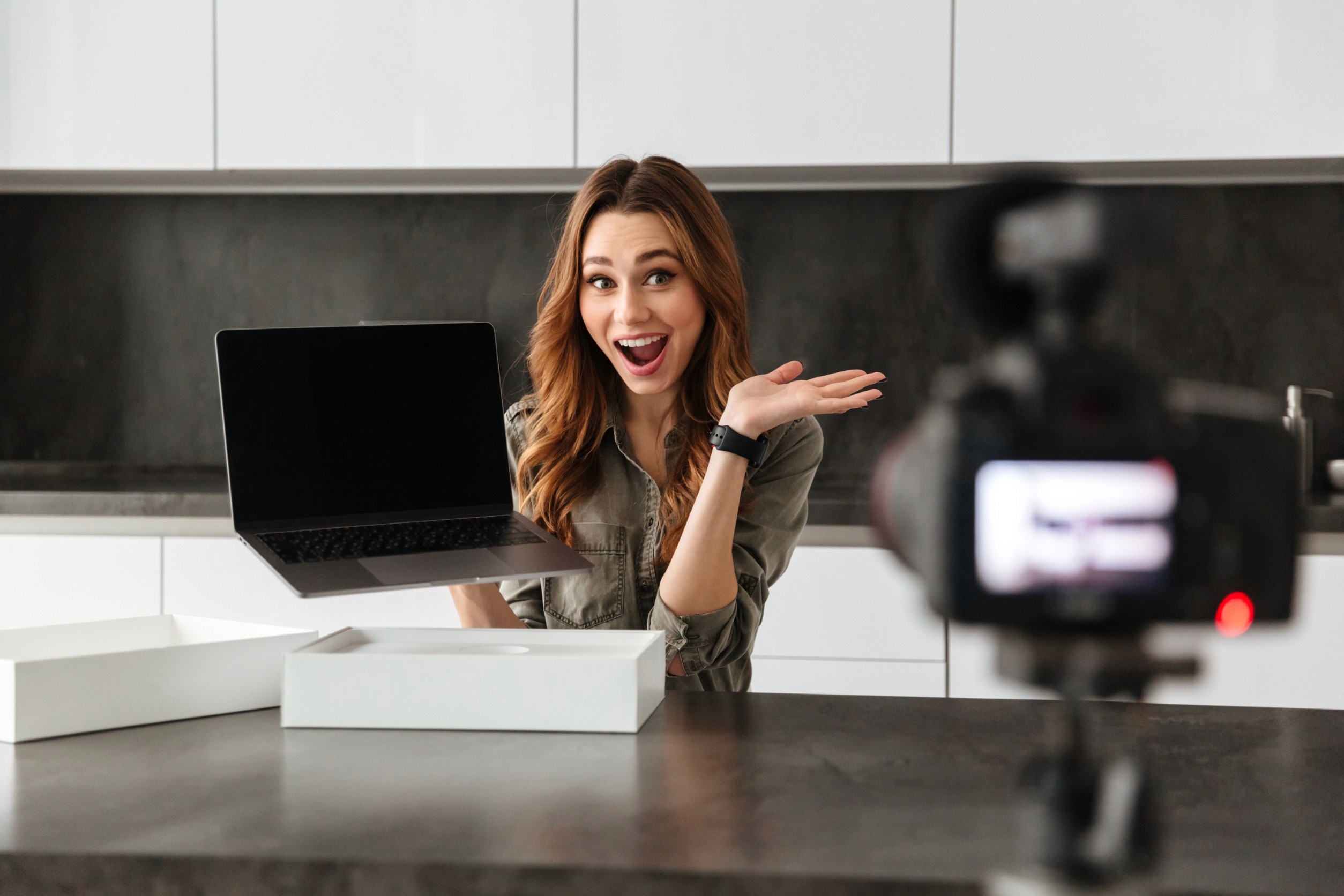 Enthusiastic young woman influencer filming a product unboxing video for a campaign, showcasing an open laptop and a white product box on a sleek kitchen counter. A digital camera records her as she excitedly gestures while holding the laptop.