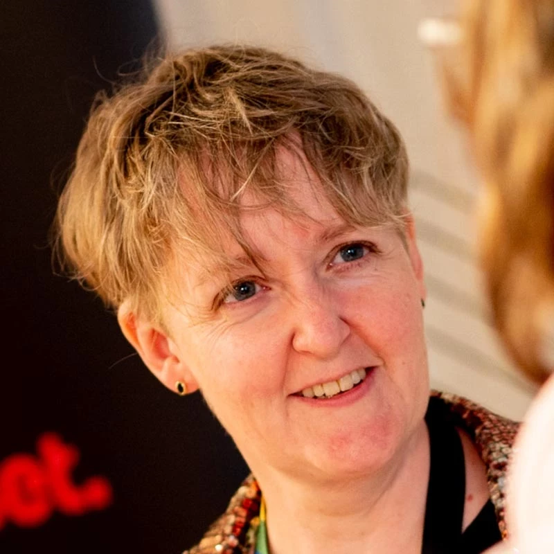 A person with short light-brown hair appears in a warmly lit indoor environment. A dark backdrop with red text and another person slightly visible add context to the scene.