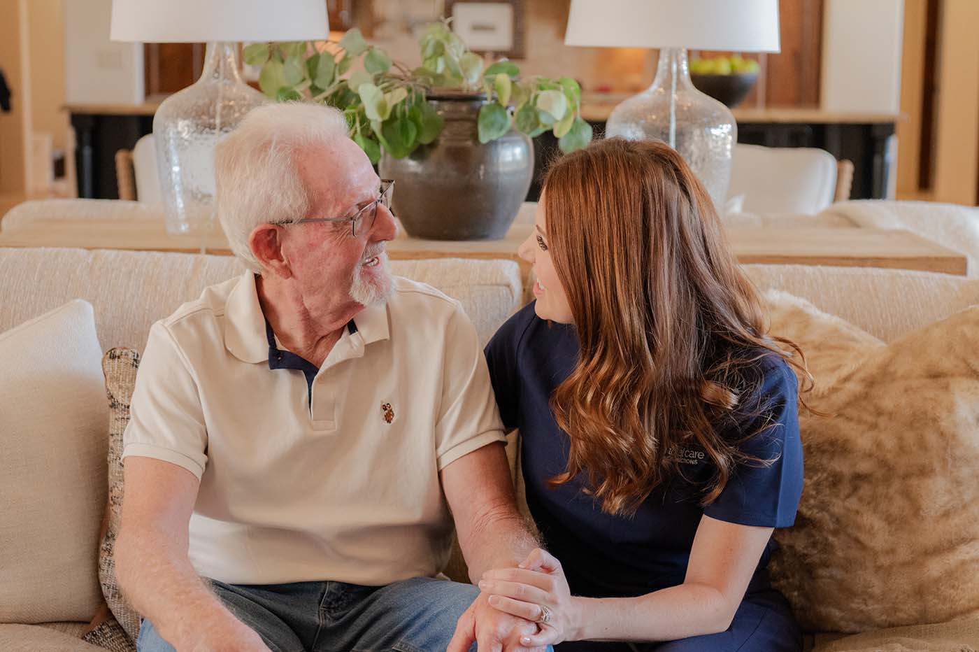 An older man and a younger woman sit on a couch, holding hands and looking at each other.