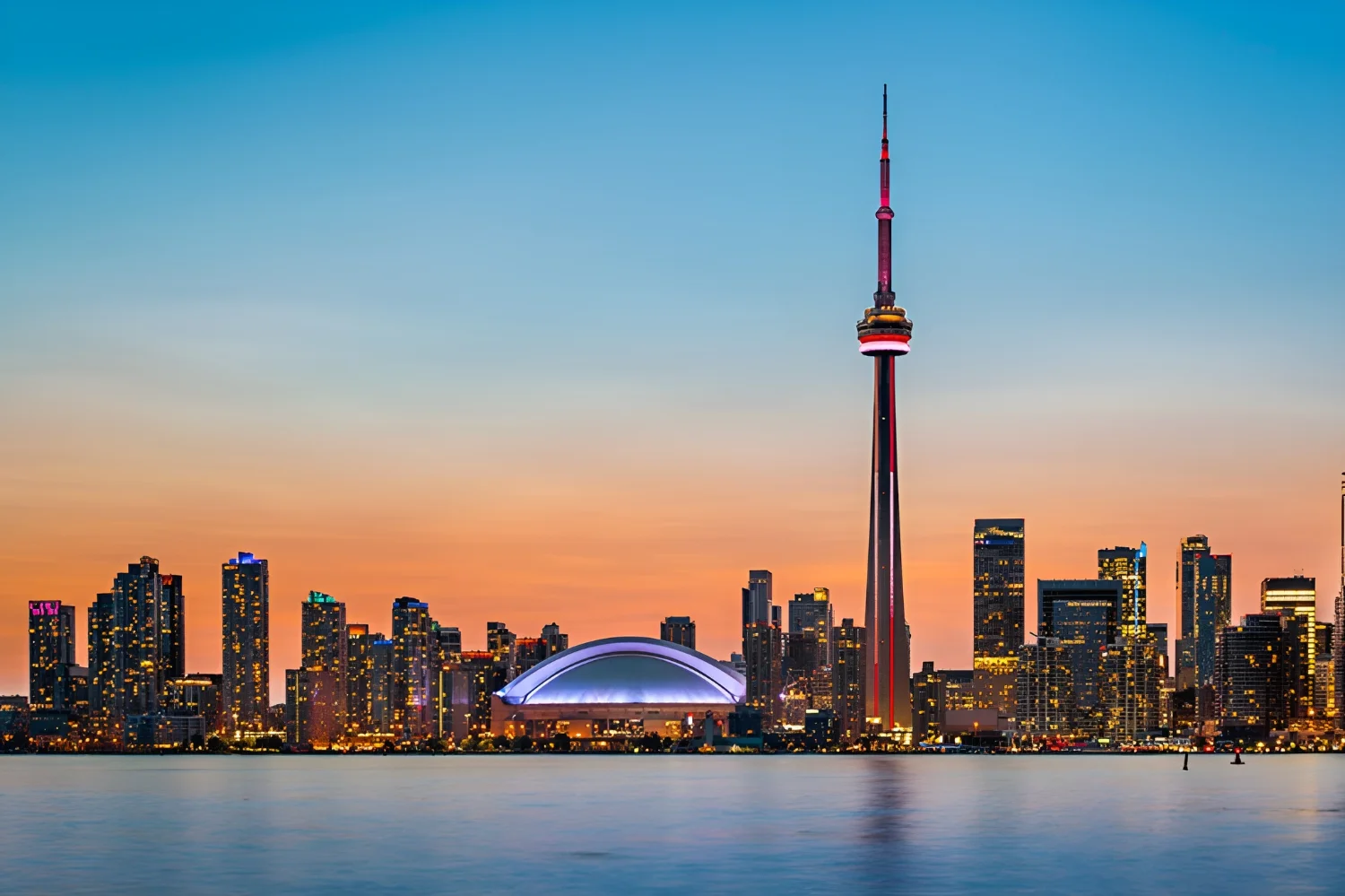 Vista do skyline de Toronto ao anoitecer, com a CN Tower iluminada e o lago Ontário em primeiro plano.
