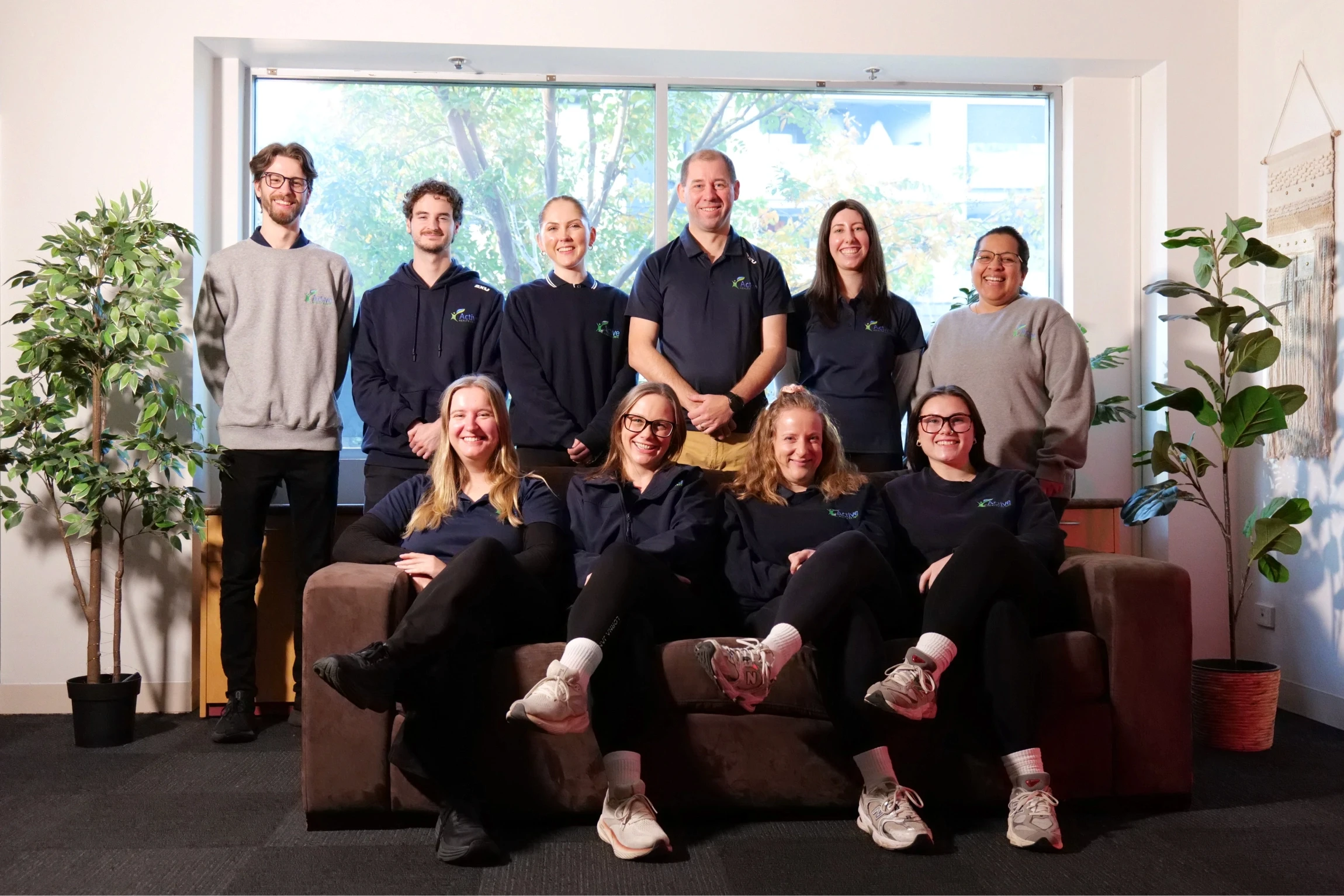 A group of ten people, dressed in casual attire including matching dark blue and gray tops, are gathered in a well-lit room, sitting and standing around a brown sofa, with large windows and green plants in the background.