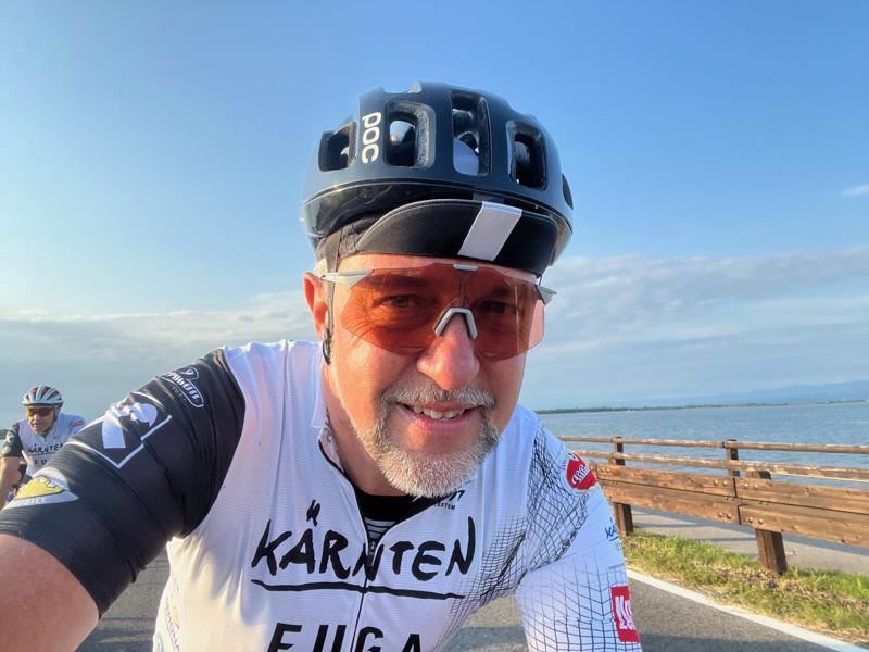 A cyclist wearing a black helmet, orange-tinted sunglasses, and a white cycling jersey smiles while riding along a waterfront path on a clear day.