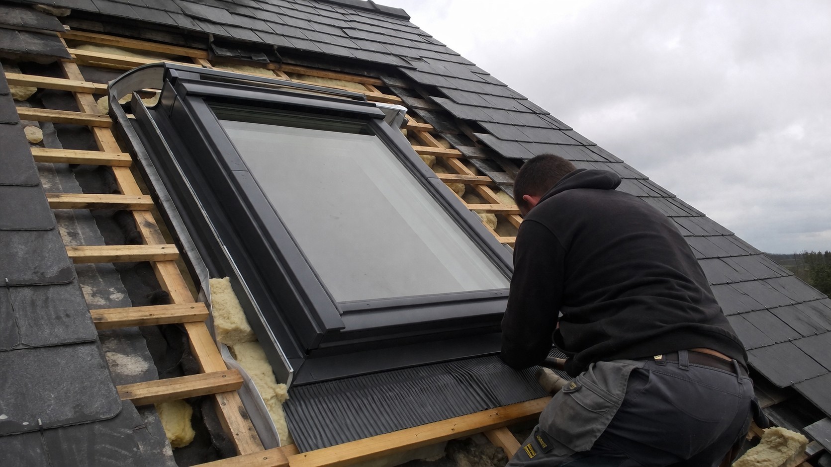 Artisan réalisant le changement d’une fenêtre de toit type Velux sur une toiture inclinée avec dépose des ardoises, isolation visible et pose du raccord d’étanchéité