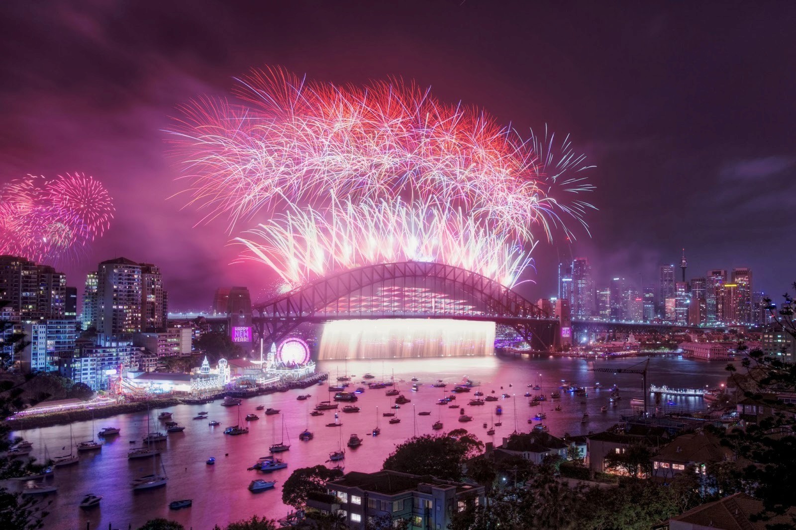 New Year's Eve fireworks explode over Sydney Harbour at night, captured with a long exposure. Boats on the water and lit-up skyscrapers in the background.