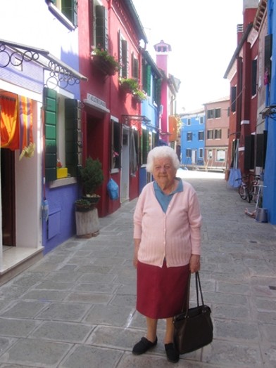 A photo of an old woman caring a bag while sitting in a narrow paved street near some colorful houses.
