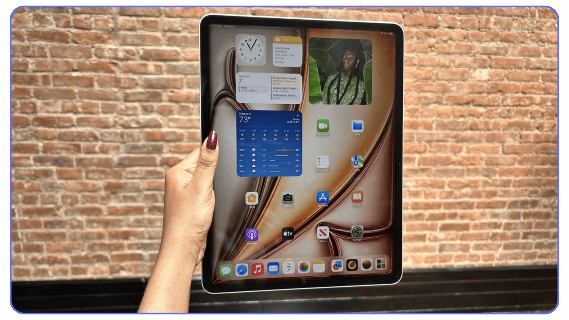 Person holding an iPad vertically in front of a brick wall, with home screen visible including clock, notes, widgets, and app icons.