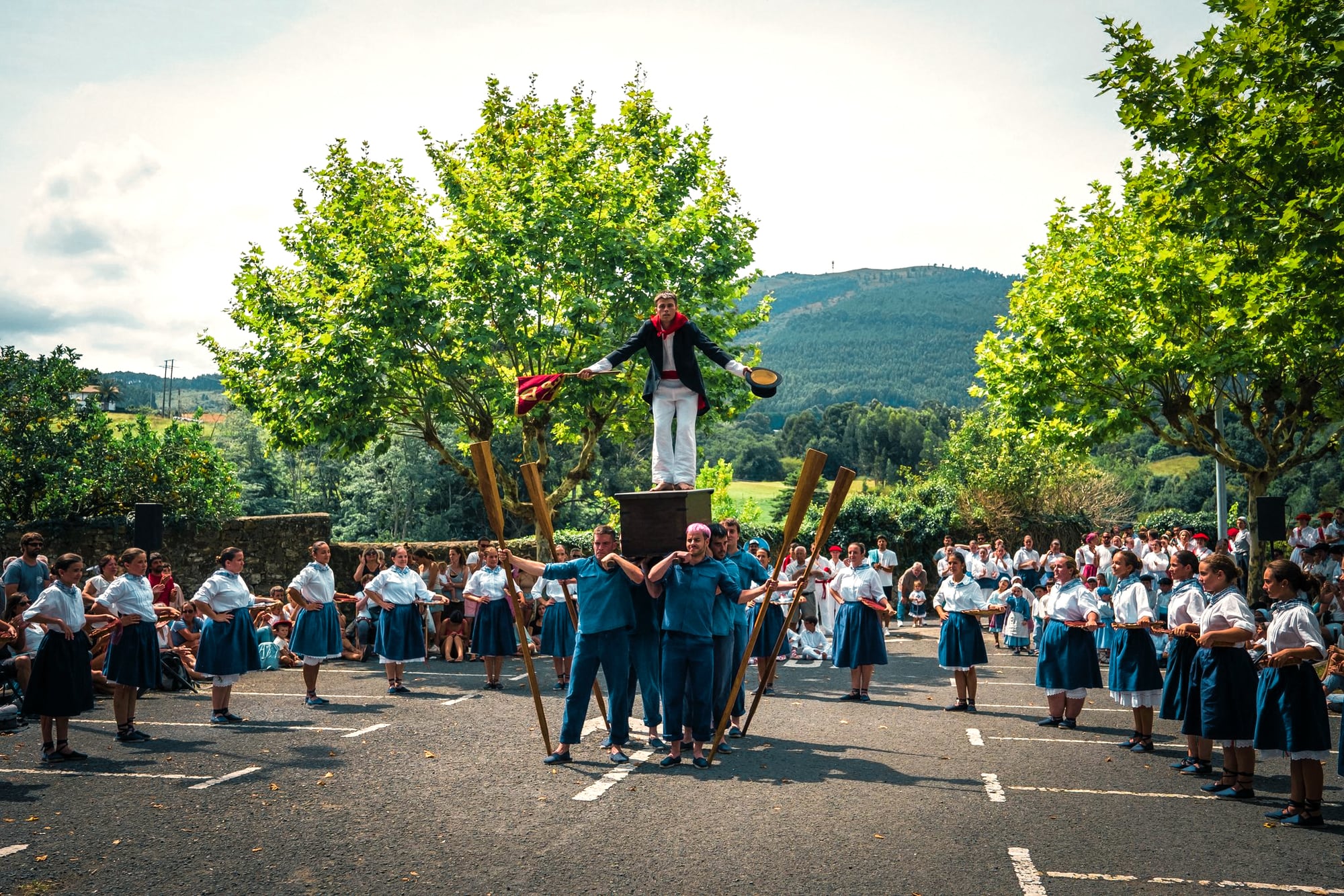 Itxas-Alde dancers performing in Bakio: a dancer stands on a box held up by the men, among oars and dancers in blue skirts