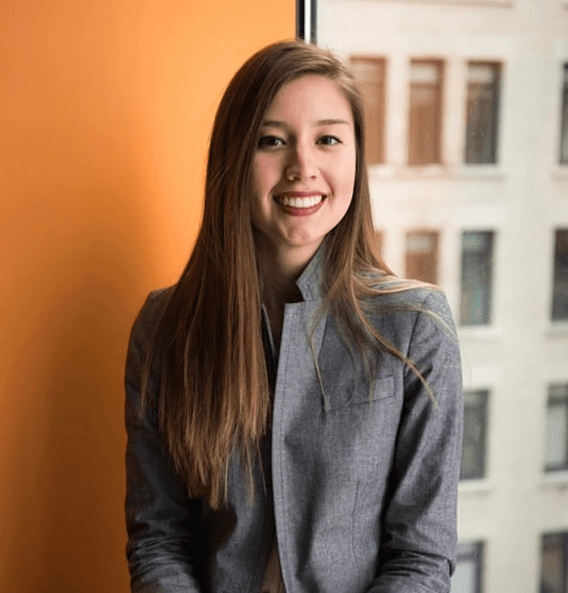Young woman with long brown hair smiles while seated in a gray suit jacket against an orange wall and large window. Urban building in background.