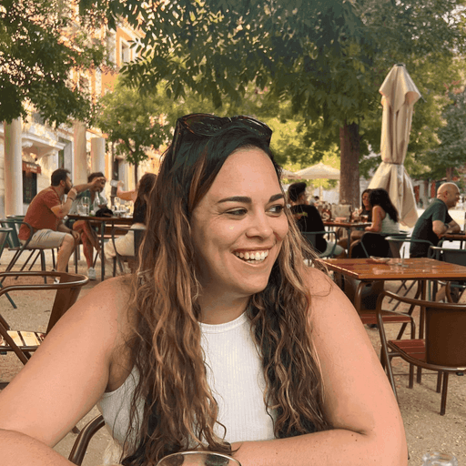 A person with long hair smiles while sitting at a café, surrounded by greenery and other patrons.