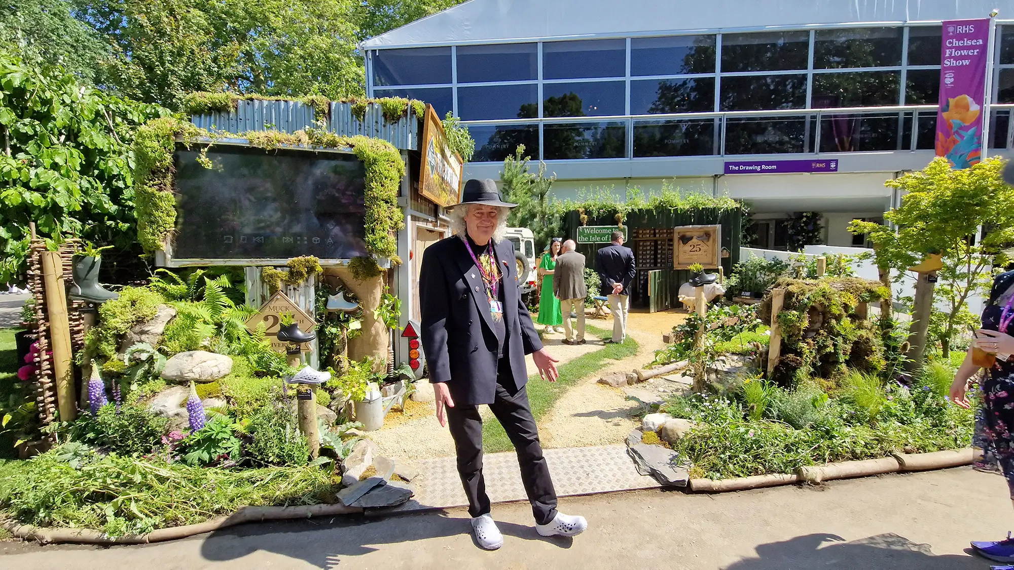 Person standing in front of a vibrant garden and building, surrounded by greenery and colorful plants.