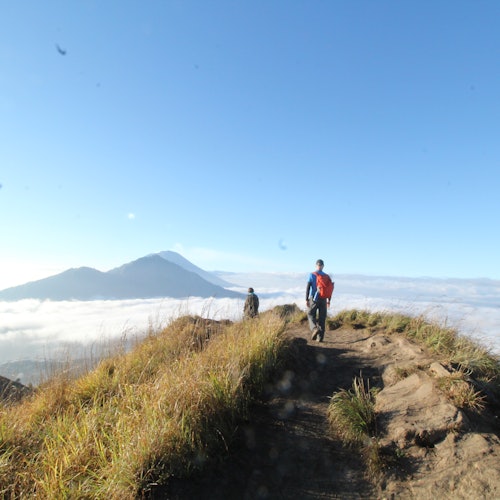 两名徒步旅行者沿着草地山顶的土路行走，背景是远处的群山和晴朗的蓝天。