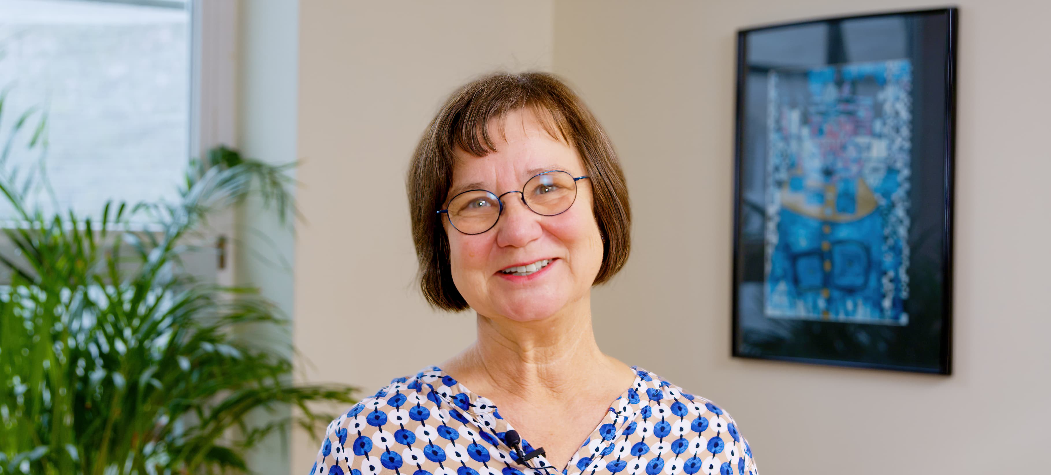 Smiling woman with glasses indoors, art and plants in background.
