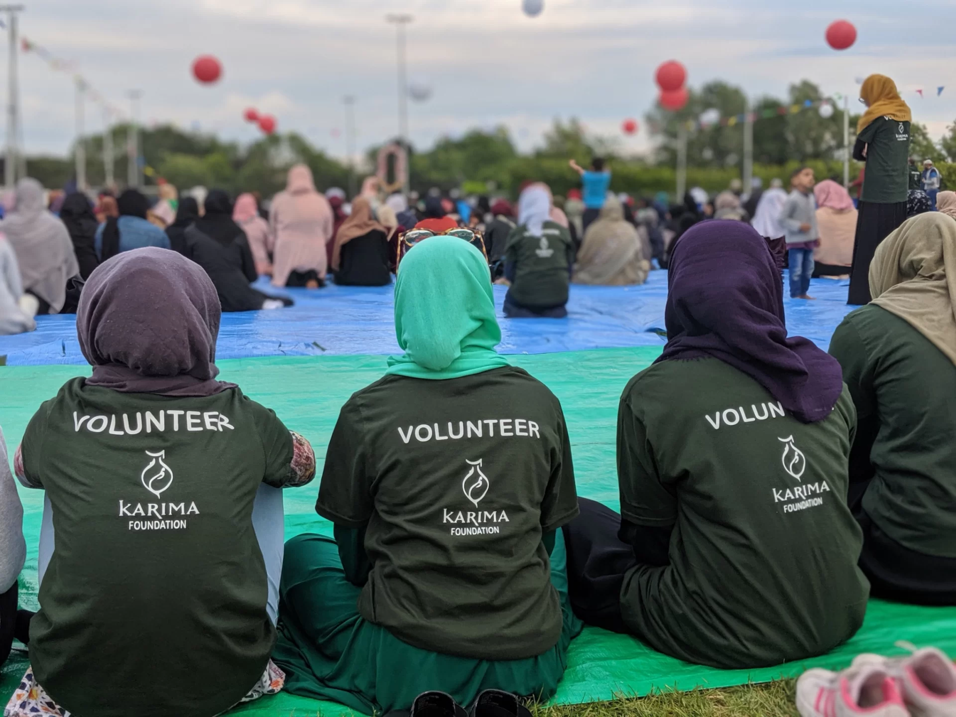 Volunteers in Karima Foundation t-shirts sitting on ground sheets at a large outdoor community gathering, with bunting and balloons overhead.