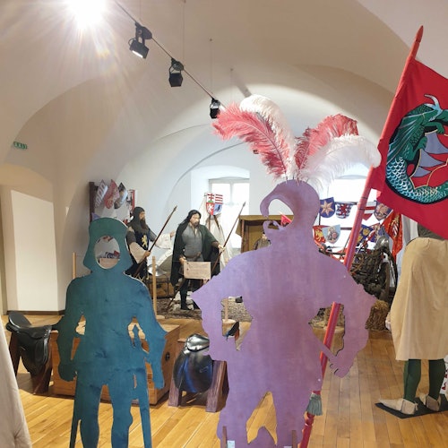 Medieval museum exhibit with knight cutouts, feather plumes, and a red dragon flag, people in the background viewing displays.