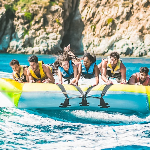 Six people in life jackets holding onto a large inflatable boat being towed on water near a rocky coastline.