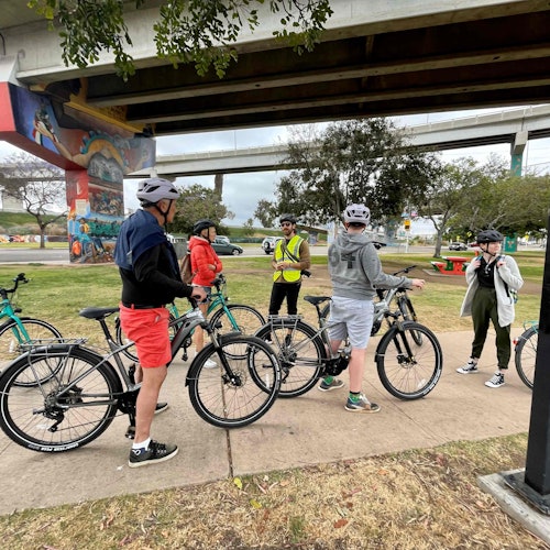 Egy csoport kerékpáros sisakot viselve gyűlik össze egy híd alatt, körülöttük kerékpárokkal és zöld növényzettel. Egy falfestmény látható a híd egyik pillérén.