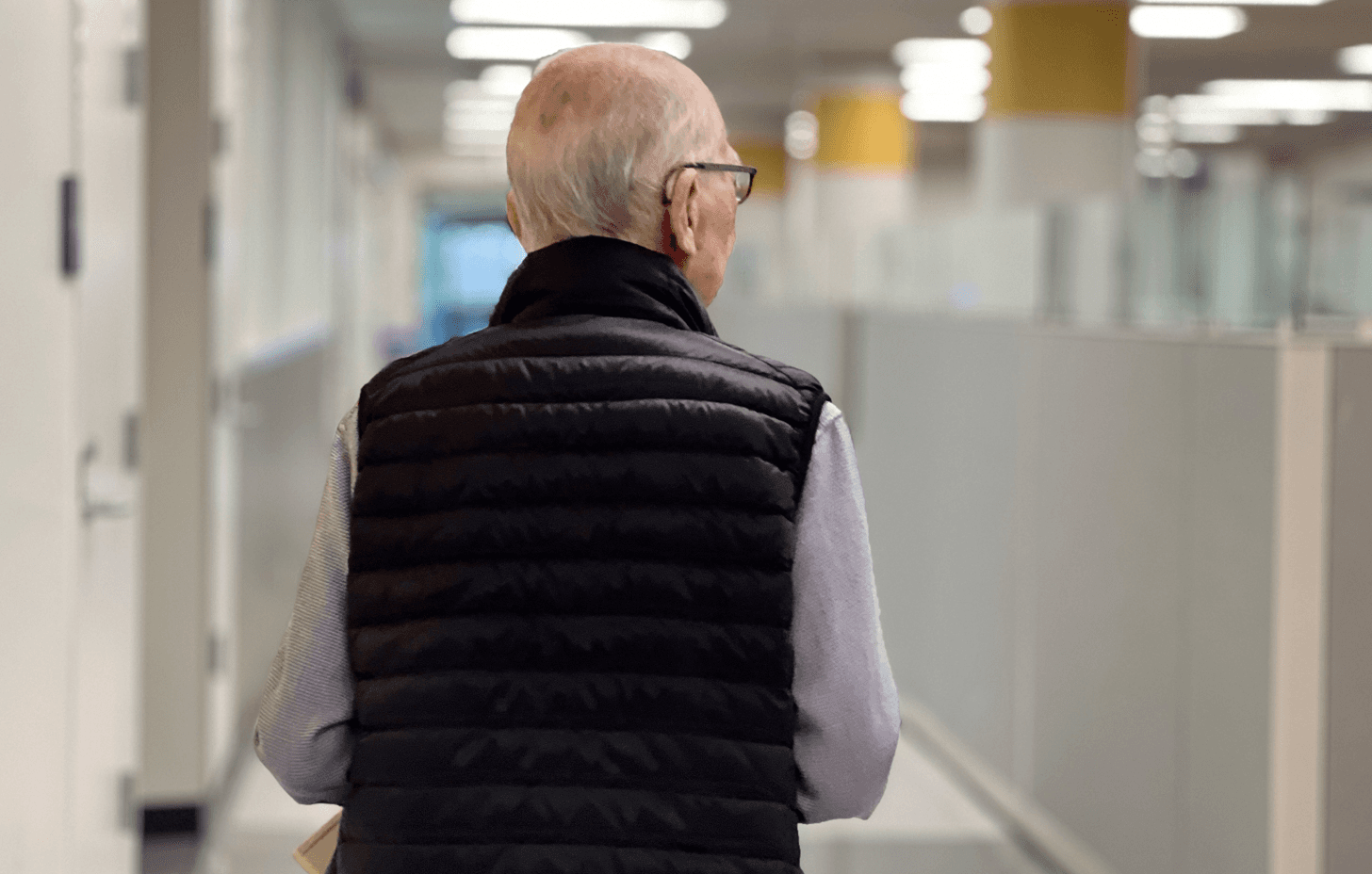 Older Professor Emeritus walks away from the camera alone down the corridor of an elite academic lab.